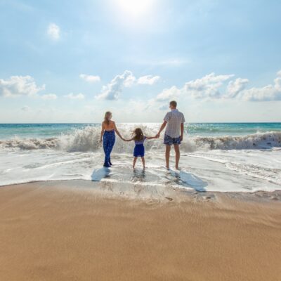 A mother, daughter and father holding hands while they stand on the beach as the tide comes in on a bright day.
