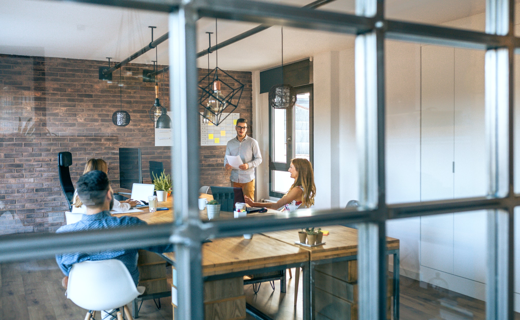 Four people are in a modern office meeting room. One person is standing and presenting, while the other three sit at a wooden table with laptops. The scene is viewed through a glass window.