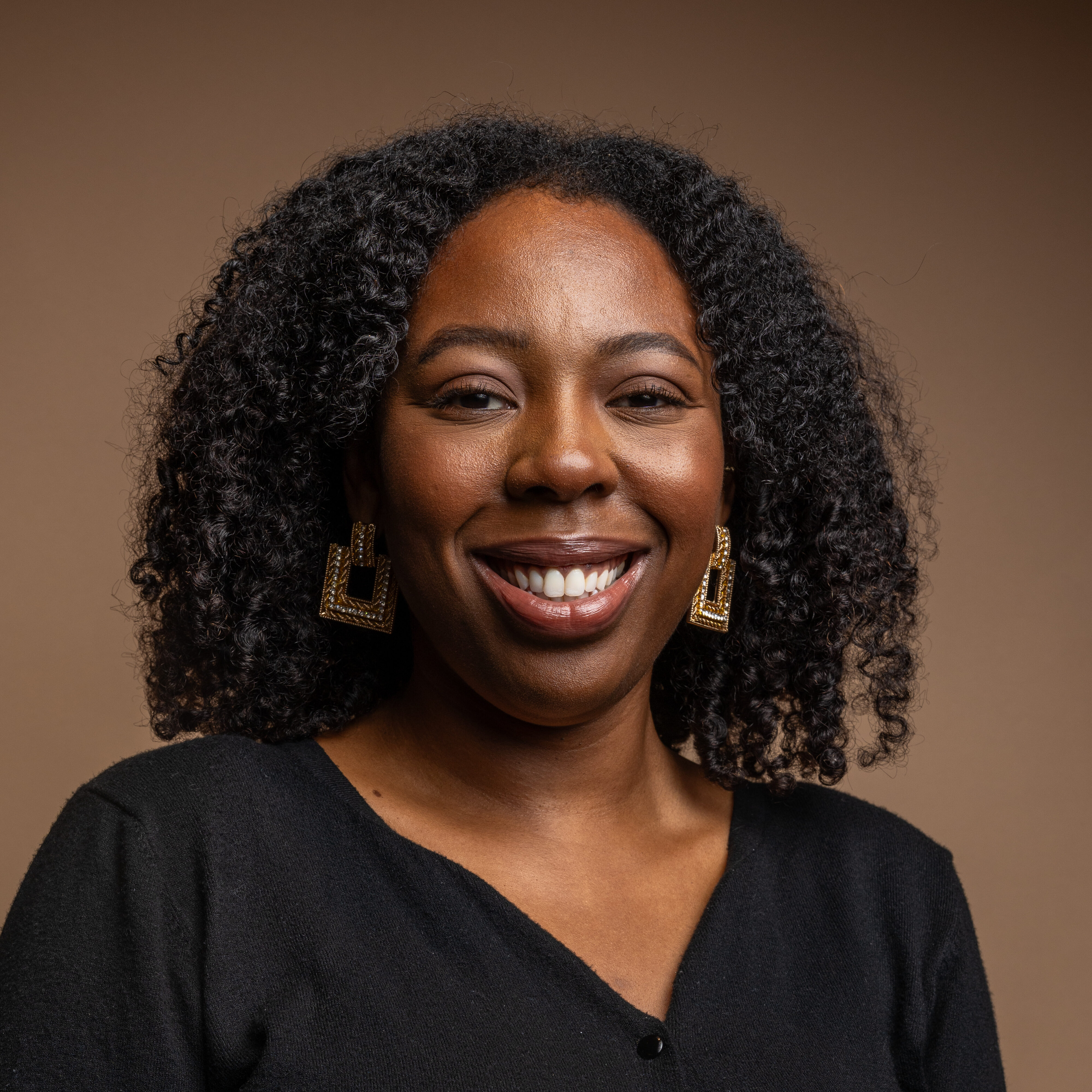 A woman with curly black hair smiles while wearing a black top and large geometric gold earrings, posed against a plain brown background.