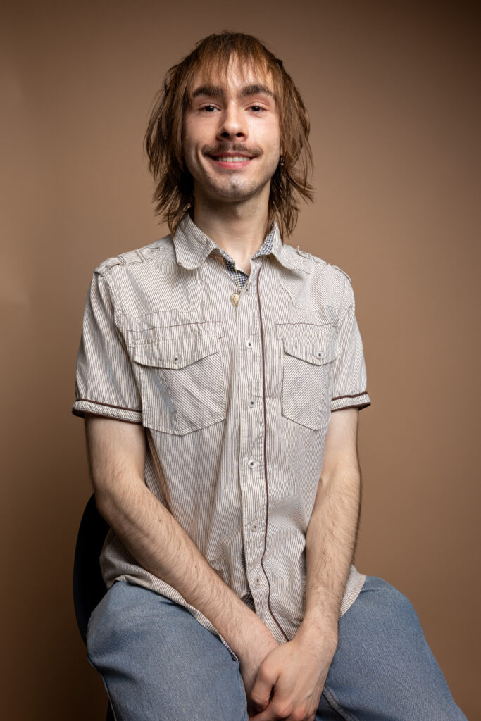 A young person with shaggy brown hair, a light mustache, and an earring smiles at the camera. They are wearing a light striped shirt and a necklace, with a plain beige background.