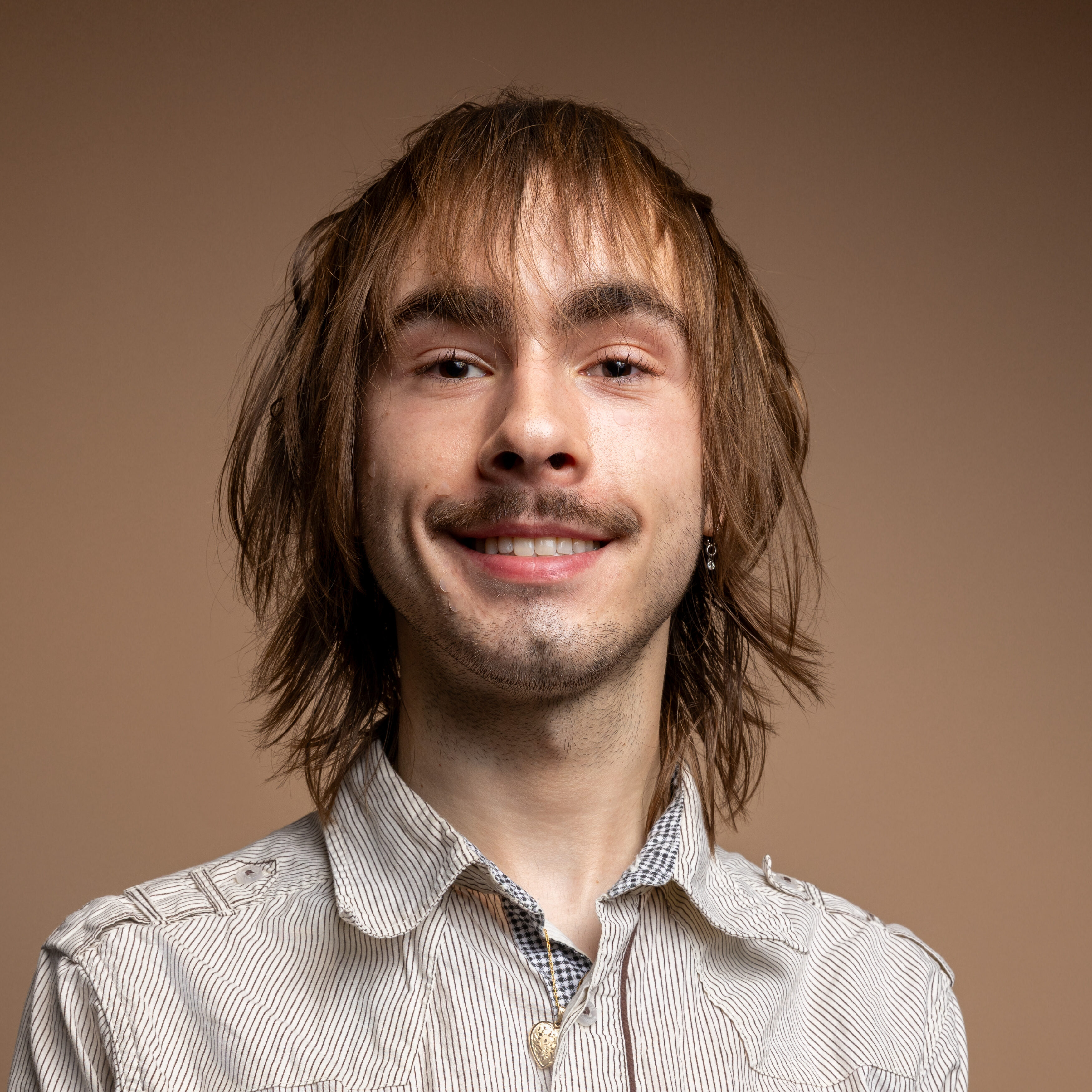 A young person with shaggy brown hair, a light mustache, and an earring smiles at the camera. They are wearing a light striped shirt and a necklace, with a plain beige background.