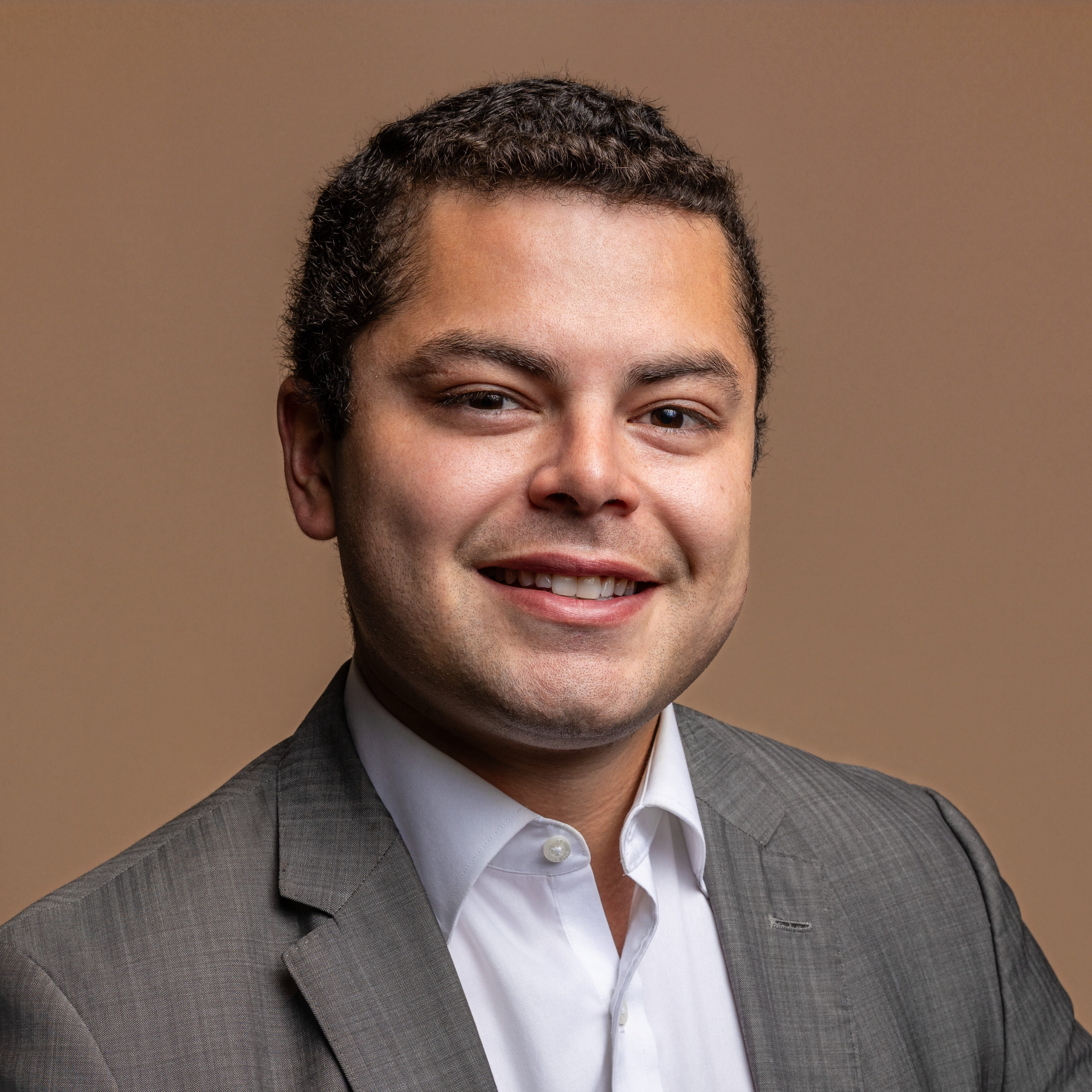 A man with short curly hair wearing a gray suit jacket and white dress shirt smiles at the camera against a plain brown background.