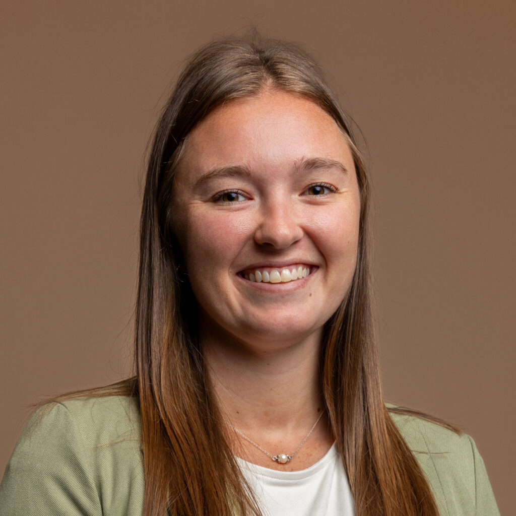 A young woman with long brown hair smiles at the camera. She is wearing a light green blazer over a white top and a delicate necklace, with a plain brown background.
