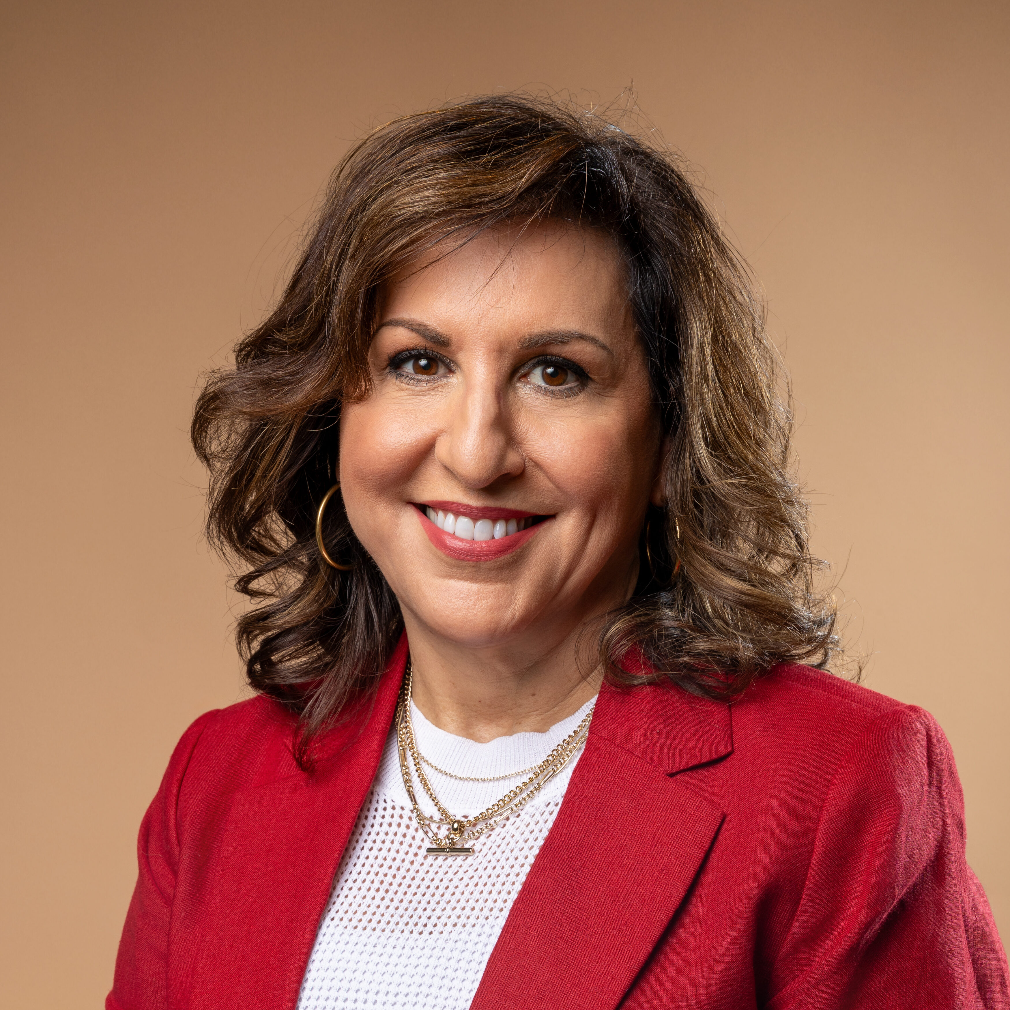 A woman with shoulder-length brown hair, wearing a red blazer over a white top, gold hoop earrings, and layered necklaces, smiles in front of a plain beige background.
