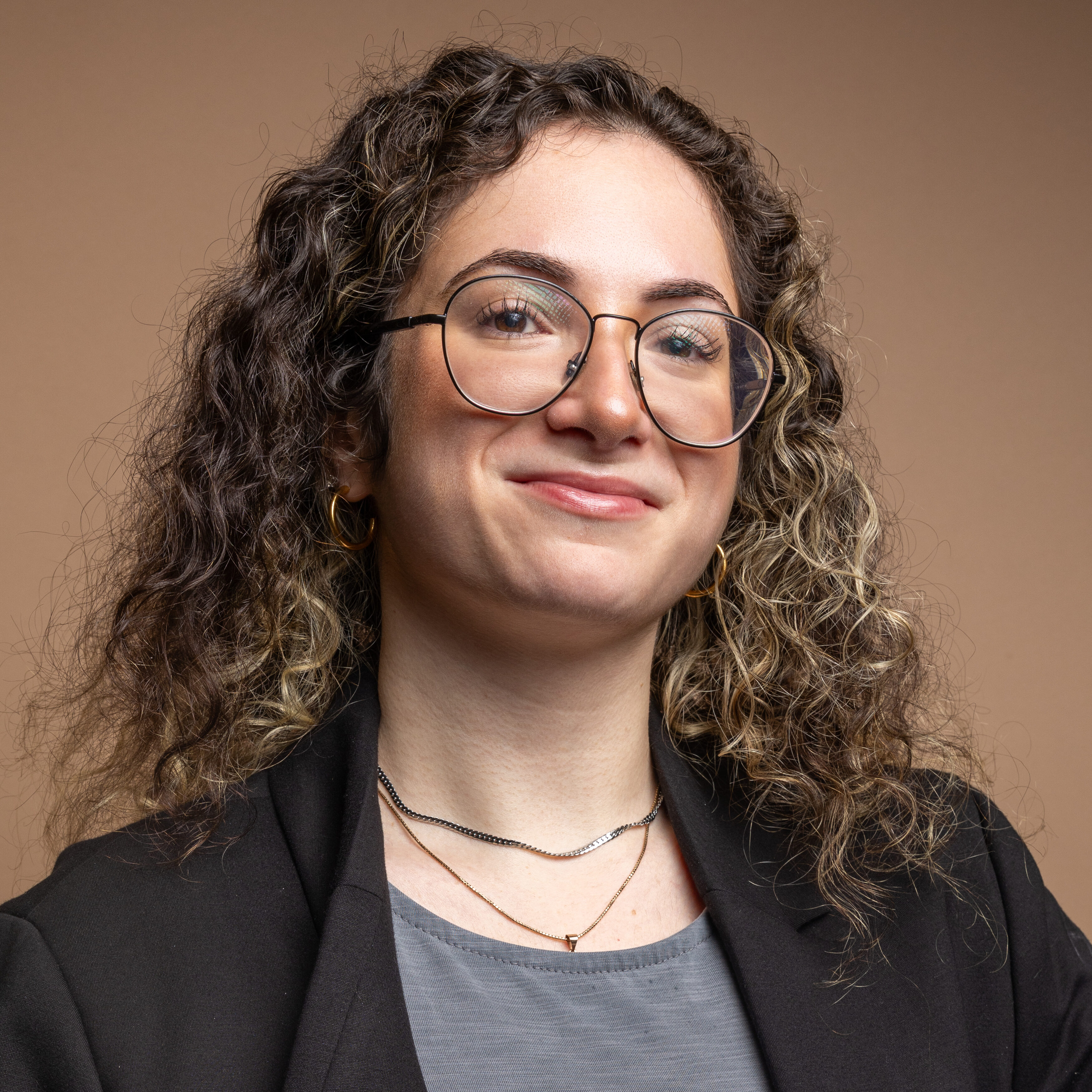 A young woman with curly brown hair and glasses smiles confidently. She is wearing a black blazer, gray top, and layered necklaces, standing against a plain brown background.
