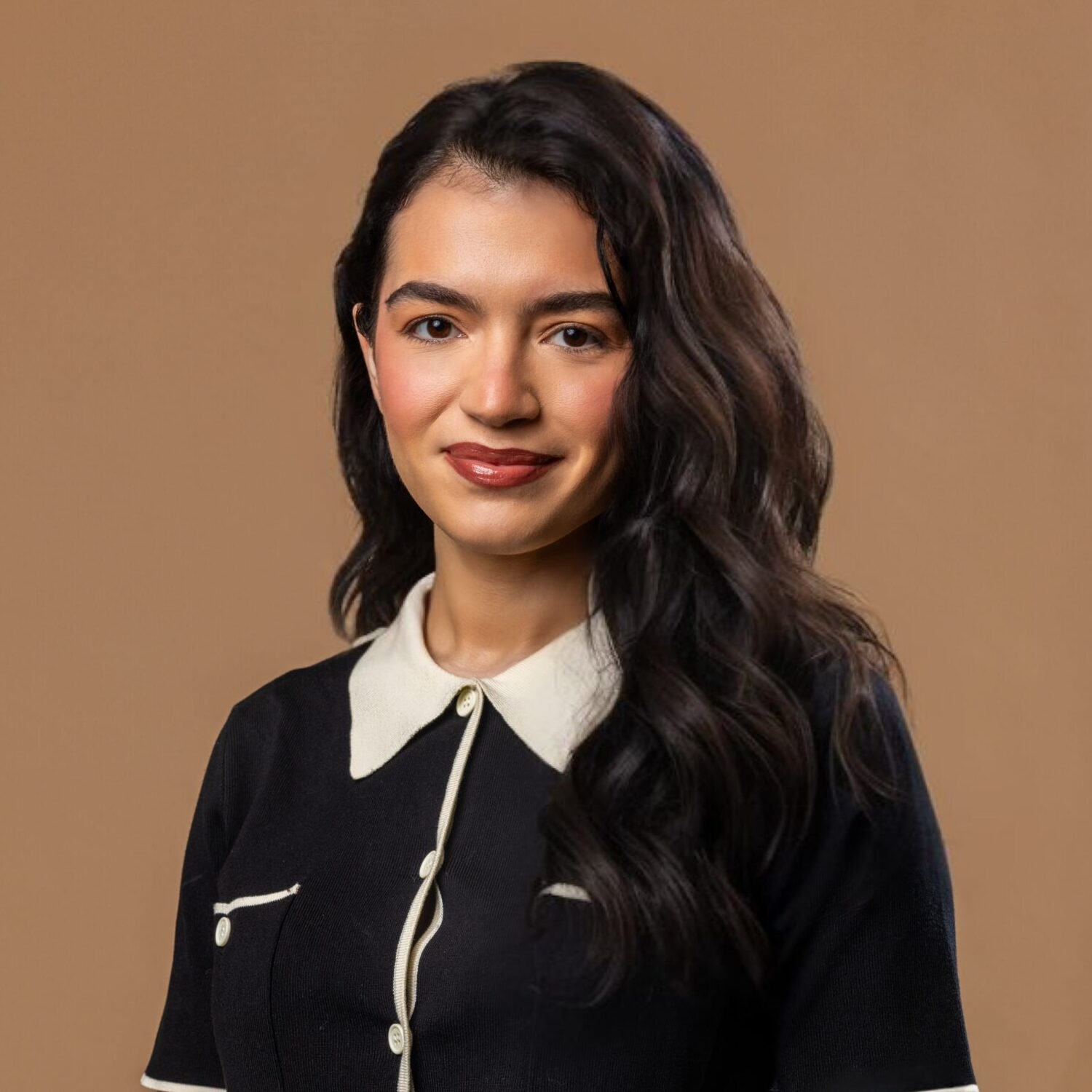 A young woman with long wavy dark hair, wearing a black dress with a white collar and buttons, smiles softly in front of a plain light brown background.