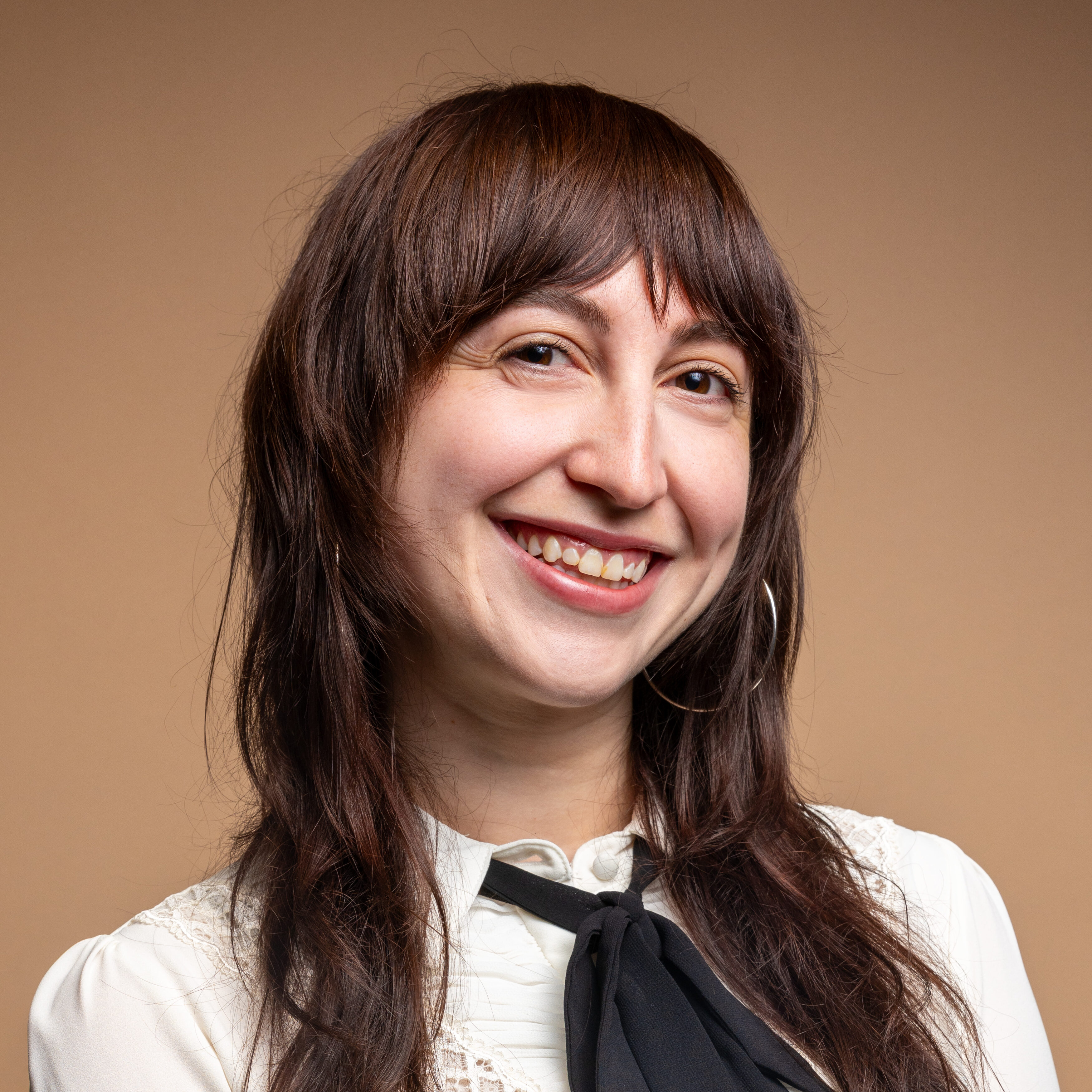 A woman with long brown hair and bangs smiles at the camera. She is wearing a white blouse with lace details and a black bow tie, standing against a plain beige background.