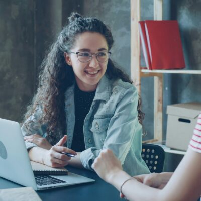 Two women are sitting at a desk having a conversation. One is smiling and holding a pen, with a laptop open in front of her. Books and office supplies are visible in the background.