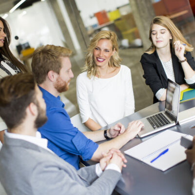 A group of five young professionals sits around a table in a modern office, engaged in discussion. One person is using a laptop while others listen and interact, with notebooks and papers on the table.