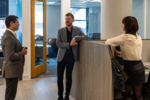 Three professionally dressed people are having a casual conversation in a modern office, standing near a cubicle and an open door. Two stand, one leans on the divider, and natural light fills the space.