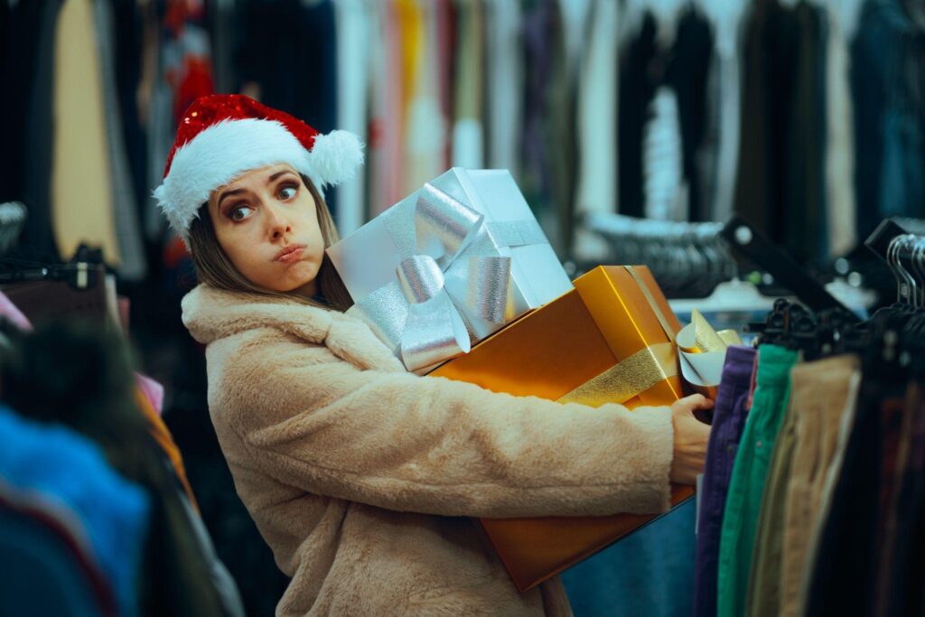 A woman wearing a Santa hat and a fur coat stands in a clothing store, holding several large gift boxes and looking overwhelmed. Racks of clothes are visible in the background.