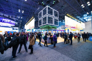 A busy convention hall with many people networking and visiting colorful exhibitor booths under bright lights and a large illuminated cube structure hanging from the ceiling.