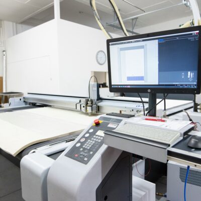 A large industrial cutting machine in a workshop, with a computer monitor displaying design software, a control panel, keyboard, and measuring tape nearby. The workspace is clean and brightly lit.