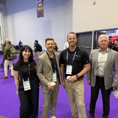 Four people stand and smile at a trade show or conference with purple carpet. They wear business attire and name badges. Other attendees and booths are visible in the background.