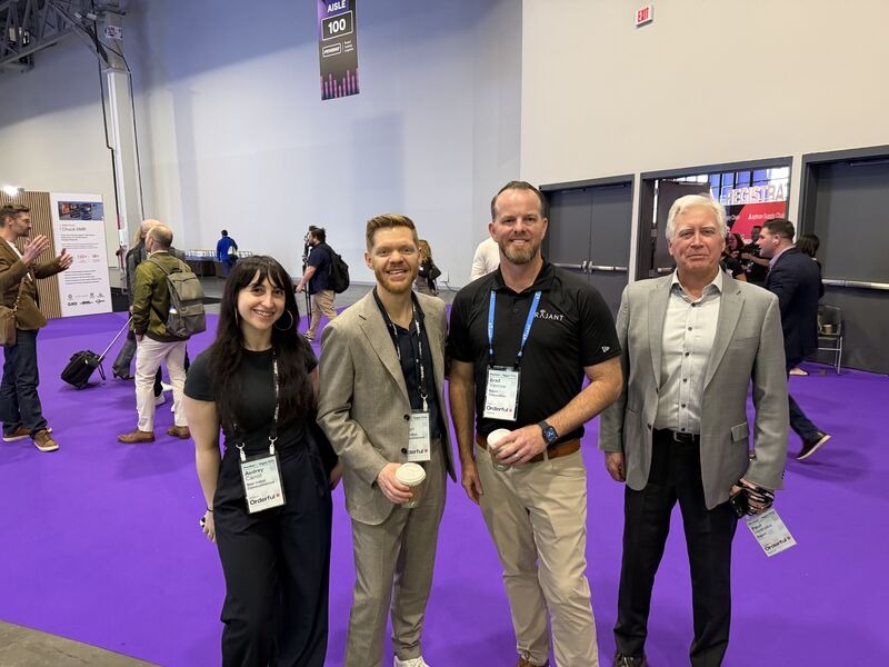 Four people stand and smile at a trade show or conference with purple carpet. They wear business attire and name badges. Other attendees and booths are visible in the background.