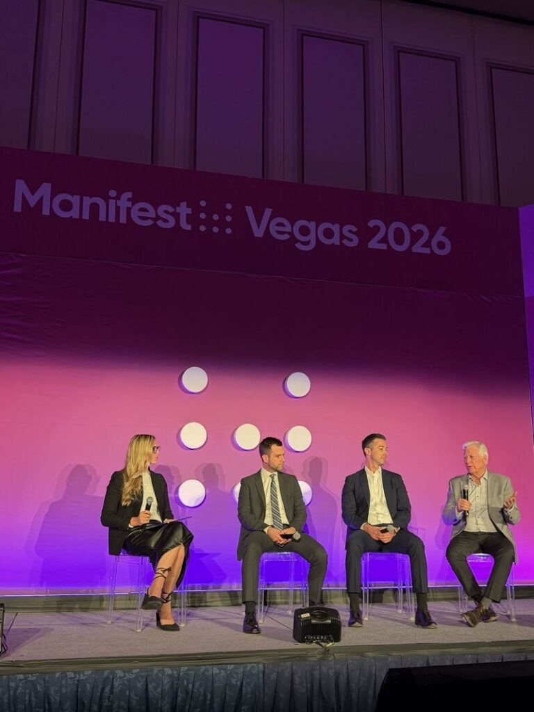 Four people in business attire sit on a stage beneath a "Manifest Vegas 2026" sign, engaged in a panel discussion. The background is purple with white dots and purple lighting illuminates the scene.