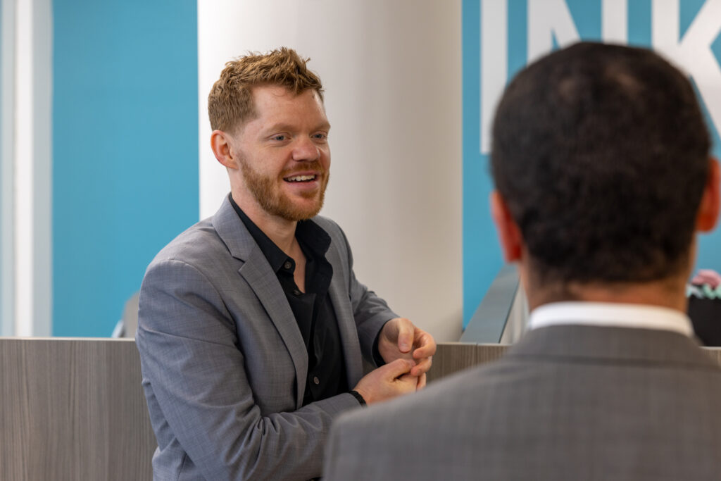 A man with reddish hair and a beard, wearing a gray suit and black shirt, smiles and gestures while talking to another man in a gray suit, seen from behind, in a modern office setting with blue walls.