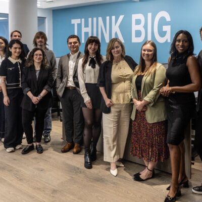 A group of fourteen professionally dressed people stand smiling in an office space in front of a blue wall that says "THINK BIG" in large white letters.