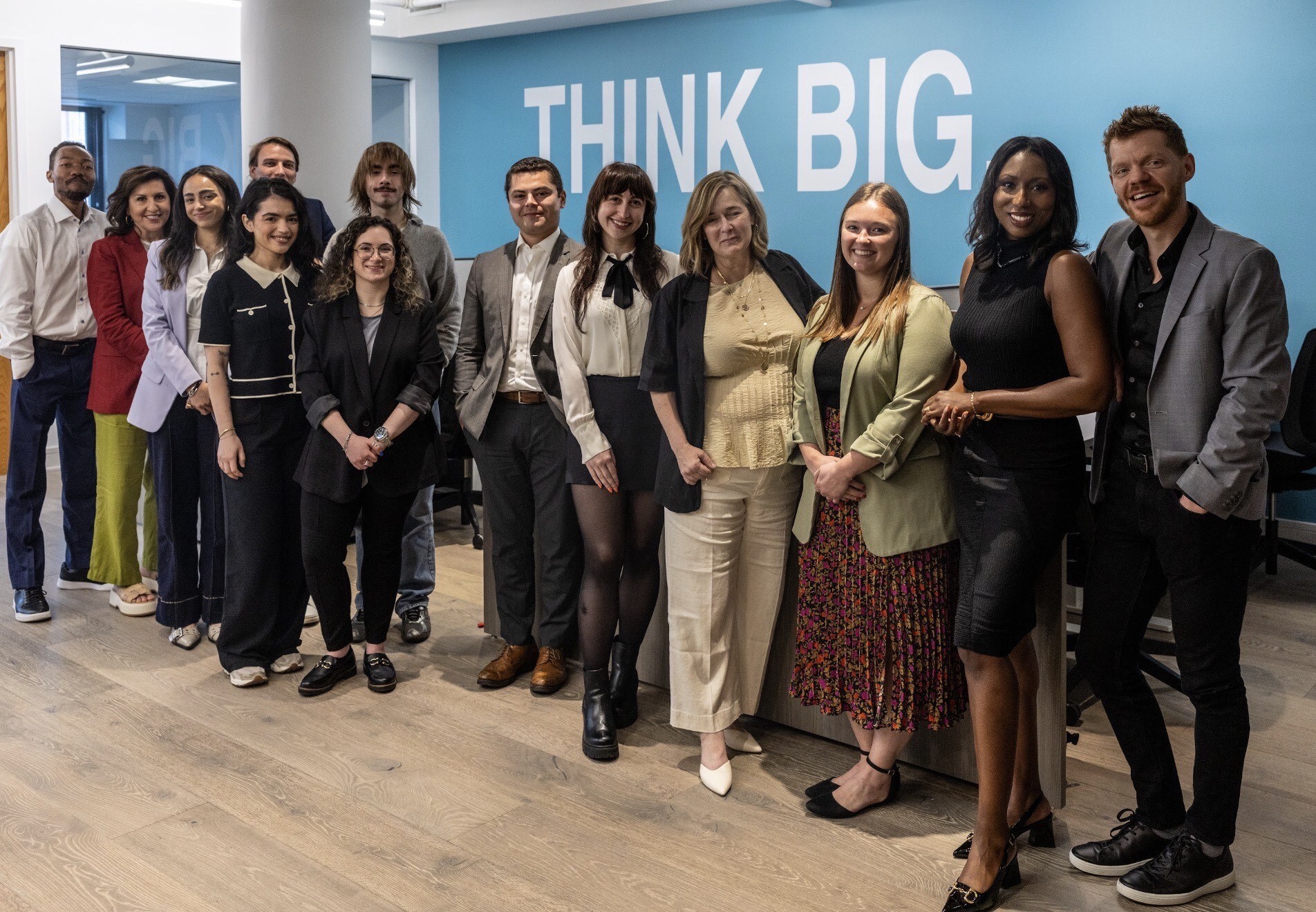 A group of fourteen professionally dressed people stand smiling in an office space in front of a blue wall that says "THINK BIG" in large white letters.