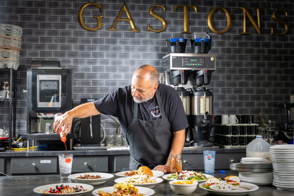A chef, skilled as an executive at a global PR agency, adds syrup to a drink behind a counter filled with plates of food in a restaurant kitchen with “GASTON'S” sign on the wall.