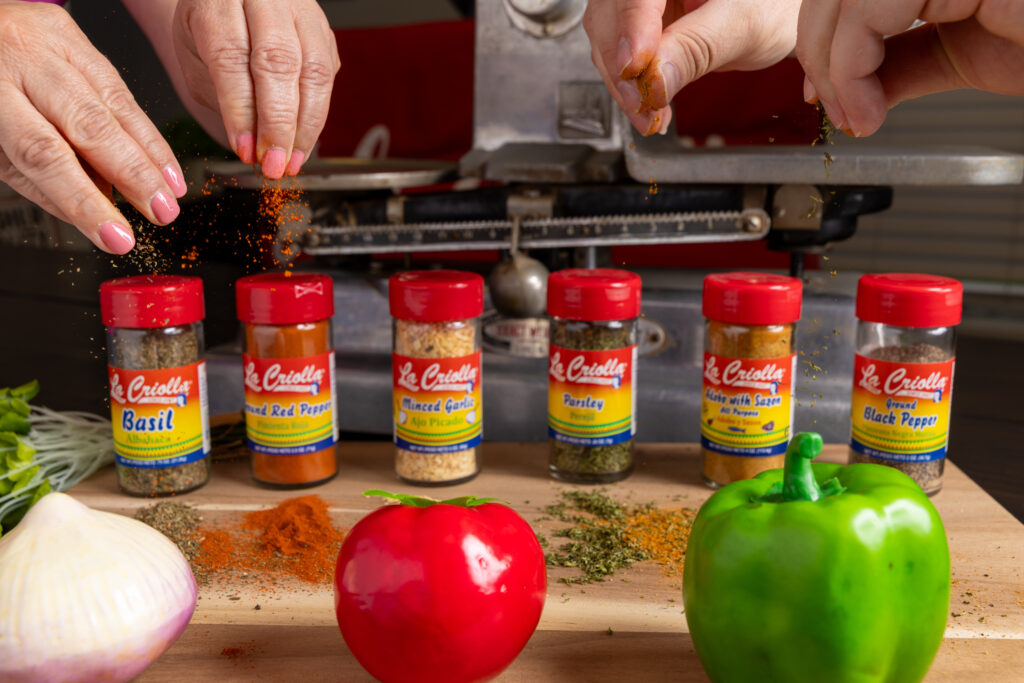 Three hands sprinkle various spices over jars labeled basil, red pepper, parsley, and black pepper—like a global PR agency bringing together diverse flavors. Fresh vegetables, including an onion and bell peppers, rest on a wooden surface.
