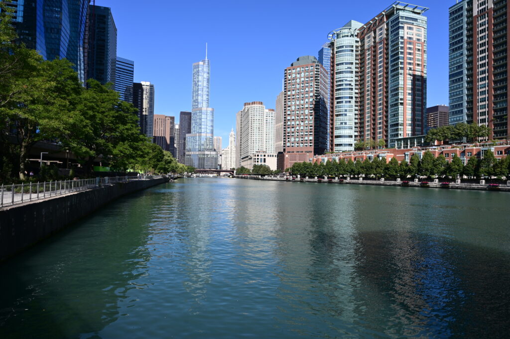 A river runs through a cityscape with tall modern buildings—home to a leading global PR agency—reflecting in the water. Trees line the left side under a clear blue sky.
