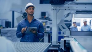A man wearing a white hard hat and blue shirt holds a tablet while inspecting machinery in an industrial factory. Two other workers in safety gear are visible in the background.