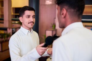A man holding a microphone interviews another man indoors. Both are dressed in white shirts, and the background features shelves and plants.