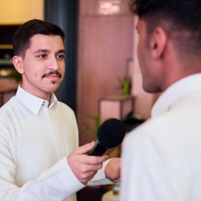 A man holding a microphone interviews another man indoors. Both are dressed in white shirts, and the background features shelves and plants.