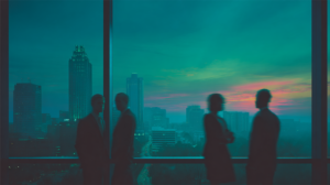 Silhouettes of four people in business attire stand and talk by large office windows overlooking a city skyline at sunset, with buildings and a colorful sky visible in the background.