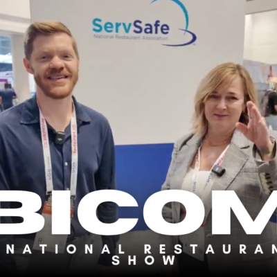 Two people stand and smile in front of a ServSafe sign at a busy indoor event. The words “BICOM @ National Restaurant Show” are overlaid at the bottom of the image.