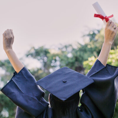 A graduate in a black cap and gown raises their arms in celebration, holding a diploma tied with a red ribbon, with trees and sky in the background.