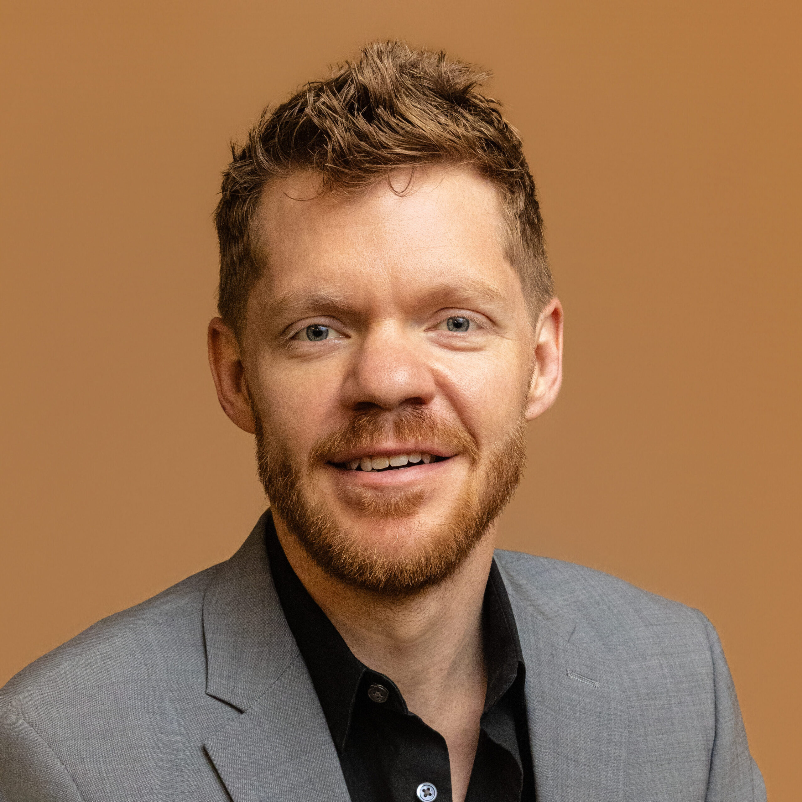 A man with short, wavy light brown hair and a beard, wearing a gray suit jacket and black shirt, smiles in front of a plain light brown background.