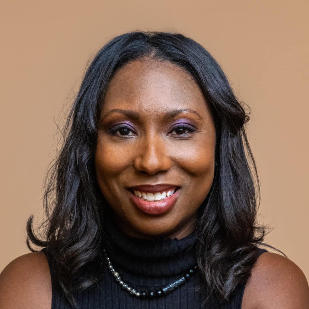 A woman with medium-dark skin, long black hair, wearing a black sleeveless turtleneck top and a beaded necklace, smiles at the camera against a plain light brown background.