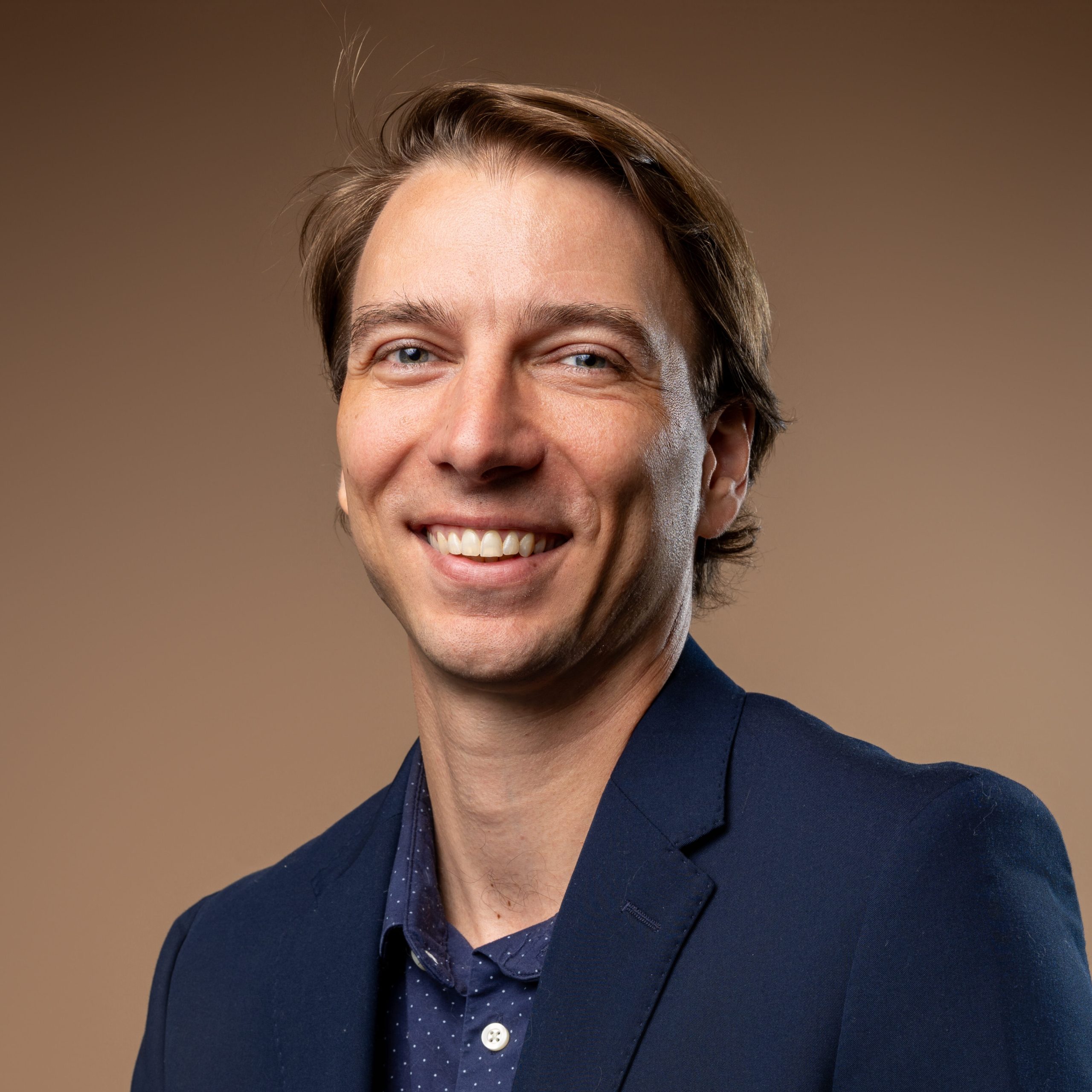 A man with light brown hair, wearing a navy blazer and a blue shirt, smiles at the camera in front of a plain brown background.