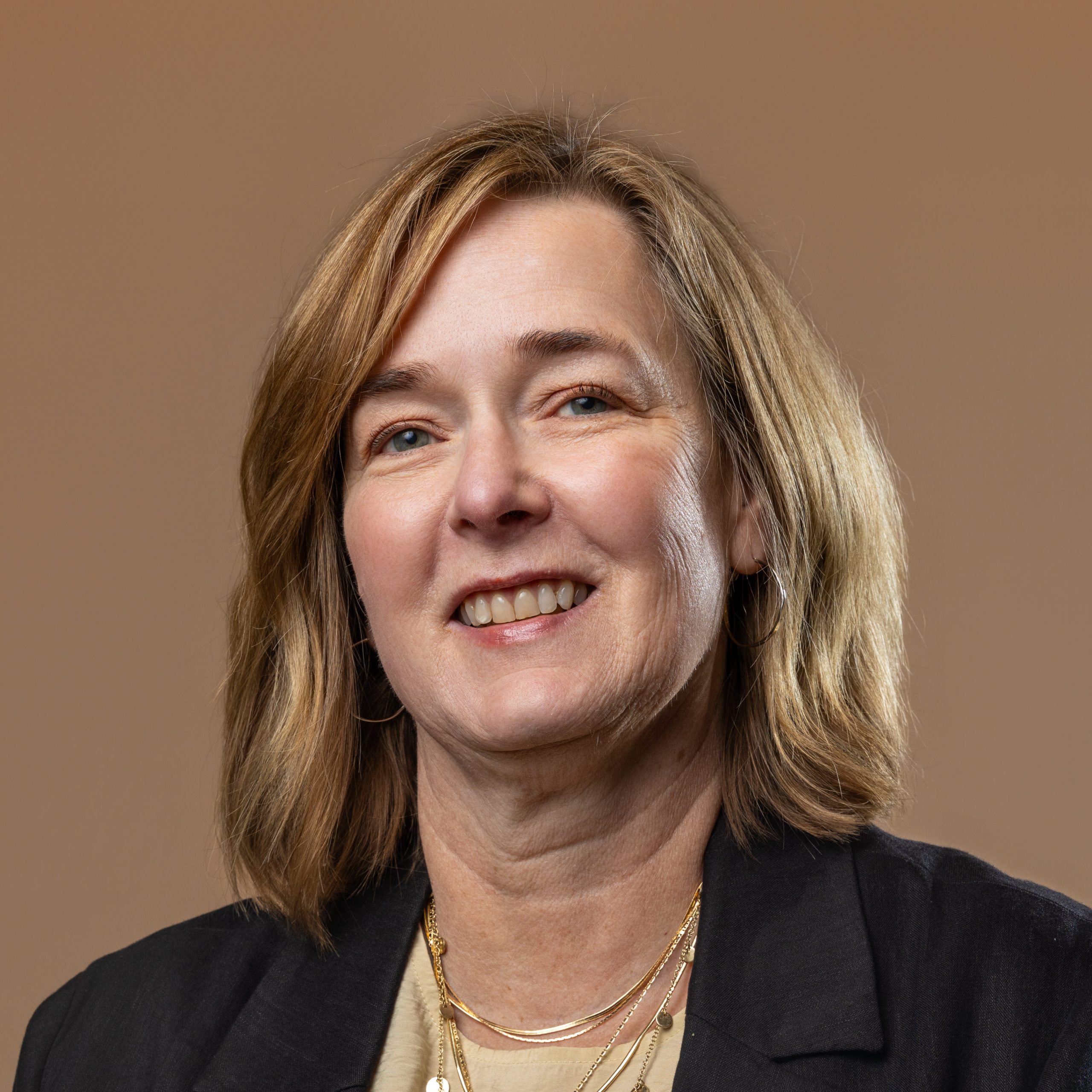 A middle-aged woman with shoulder-length light brown hair, wearing a dark blazer and a beige top, smiles at the camera against a plain brown background.