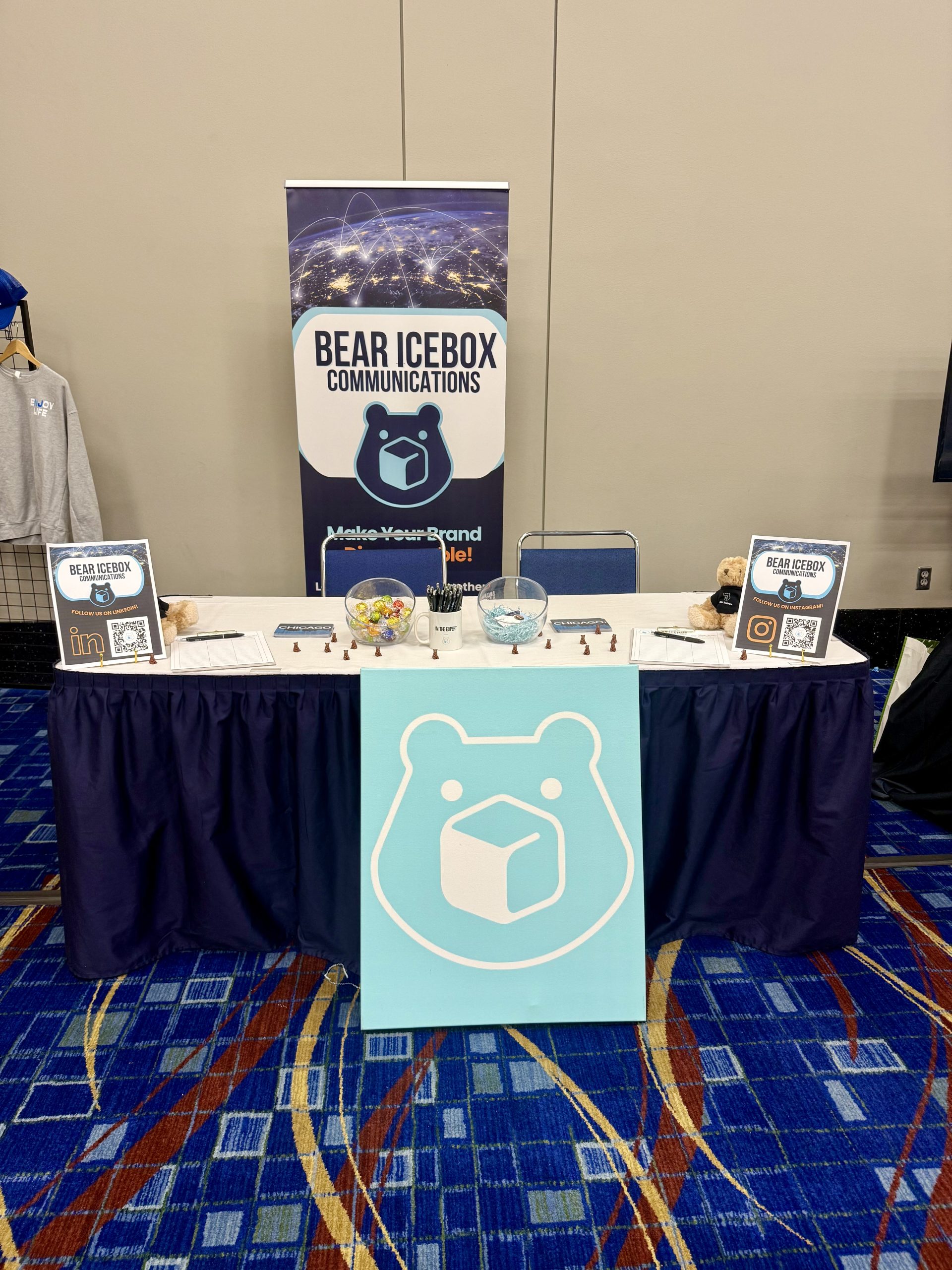 A booth with a blue bear logo and “Bear Icebox Communications” signage, two QR codes, informational flyers, a bowl of candy, pens, and business cards on a table with a dark blue tablecloth.