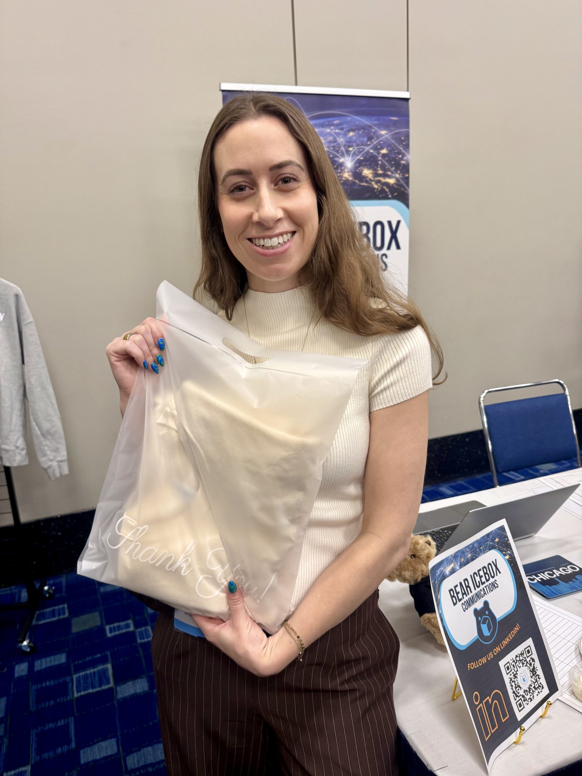 A woman smiles while holding a translucent bag with “Thank You” written on it. She stands by a table with a Bear Icebox sign, business cards, and a QR code, in a room with blue carpet and several chairs.