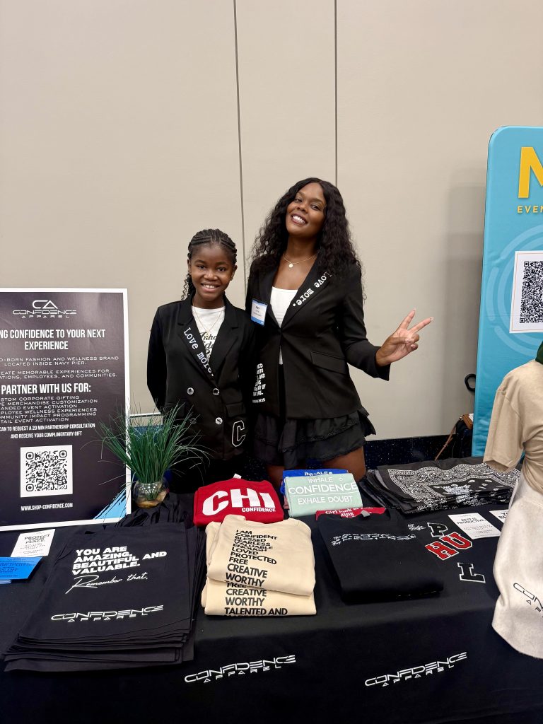 Two smiling women stand behind a table displaying shirts and promotional materials at an event. One woman flashes a peace sign. The table includes QR codes, a small plant, and signs about confidence and creativity.
