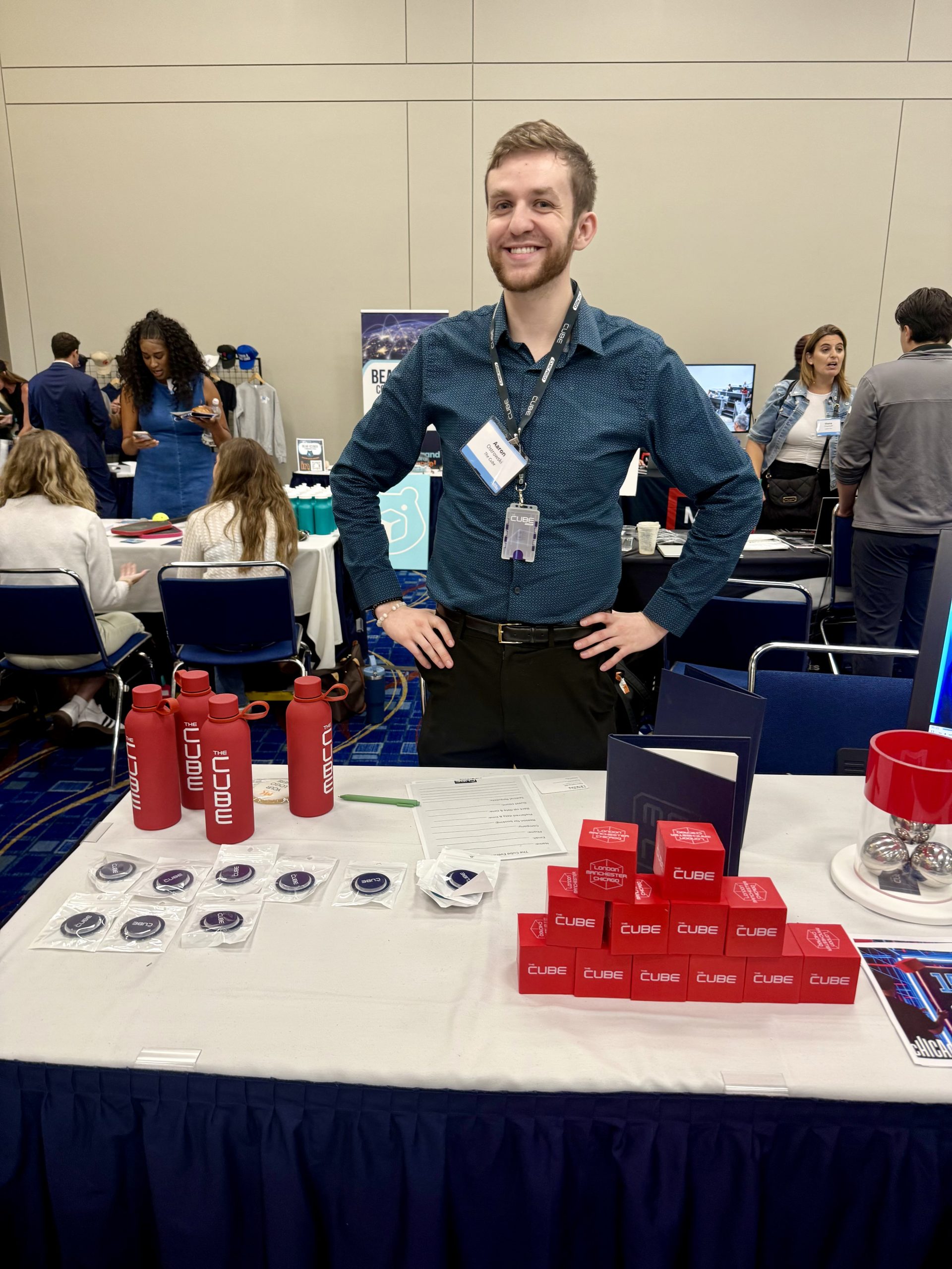 A smiling person stands behind a booth table with red Cube boxes, red water bottles, stickers, and other promotional items at a busy indoor event or expo.