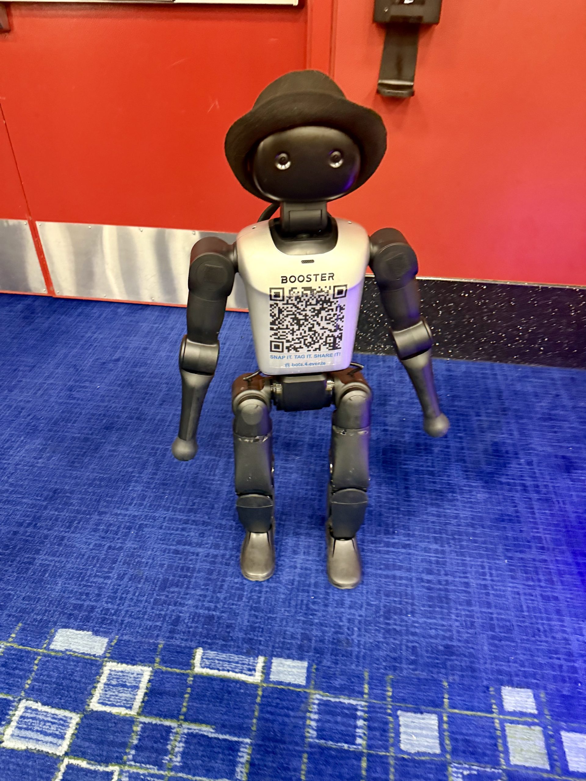 A black robot with a hat sits on a blue carpet in front of a red door. The robot has a QR code and the word “BOOSTER” on its chest.