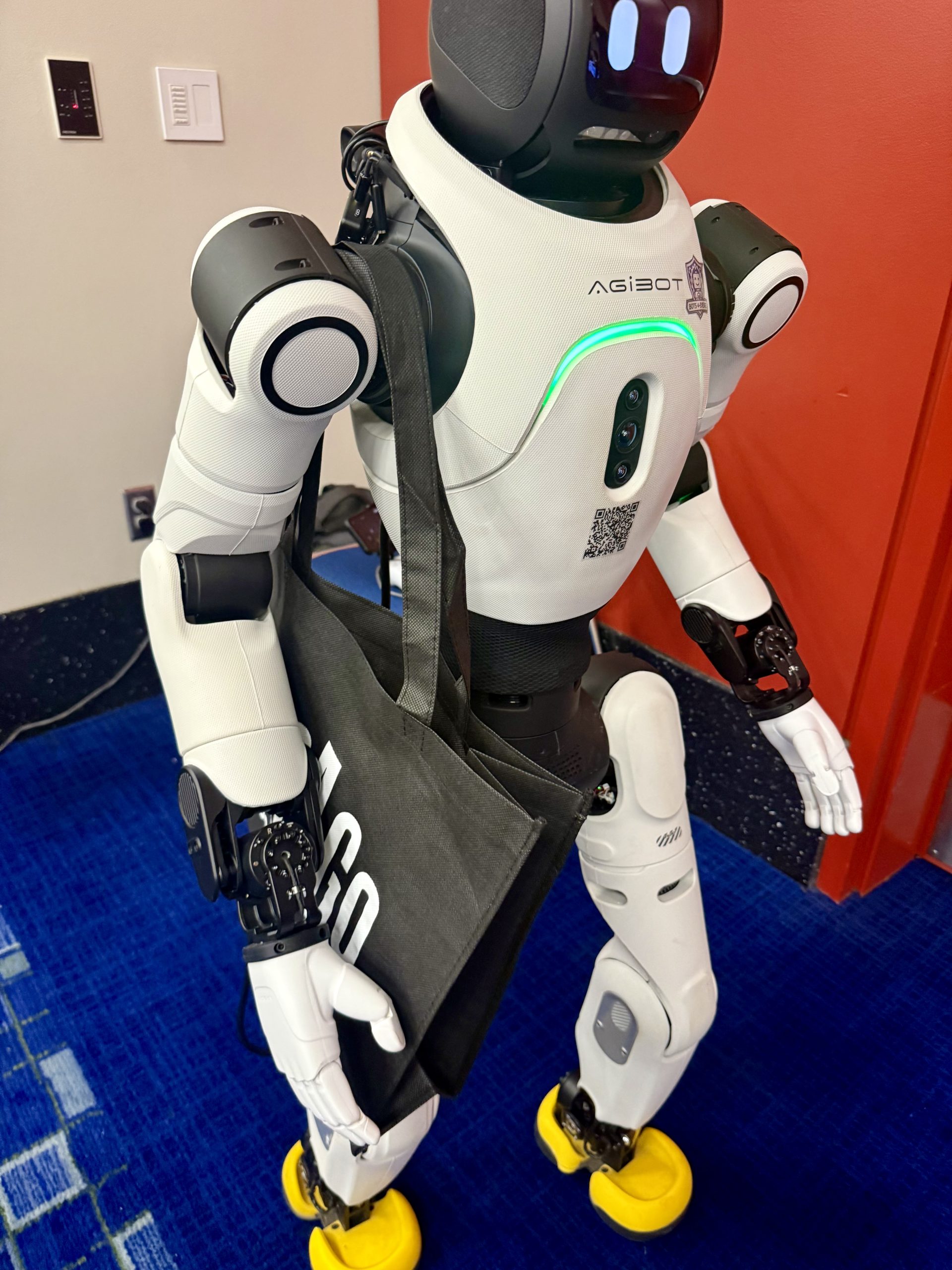 A white humanoid robot with yellow feet and a black tote bag stands on a blue carpet near a red wall. The robot has “AGIBOT” written on its chest and glowing green lights.