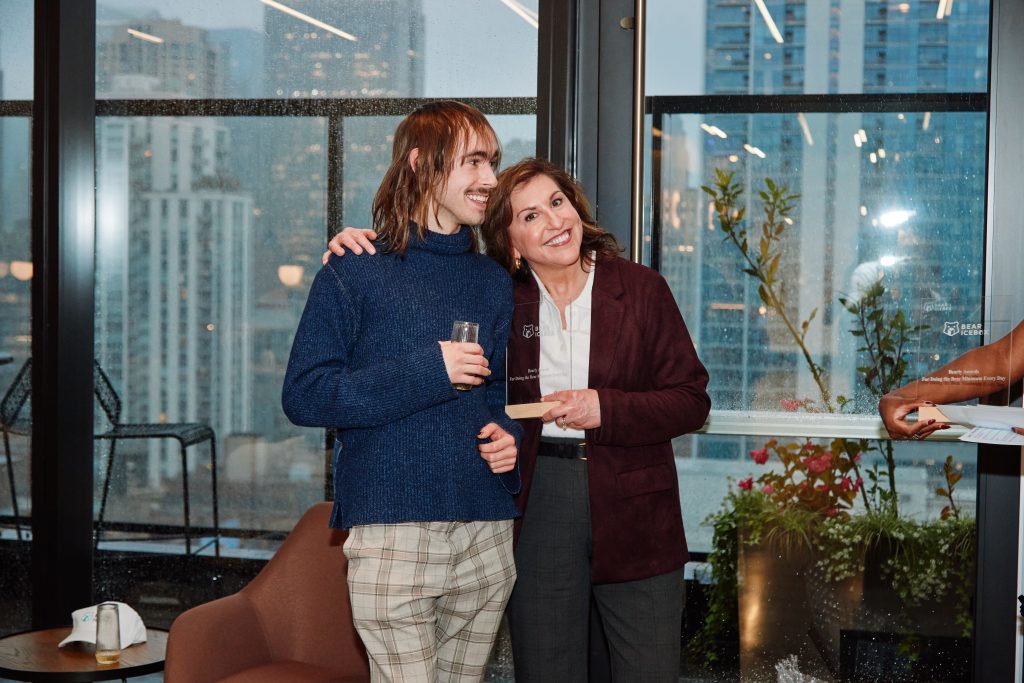 A young man and a woman stand side-by-side posing for a photo in front of a floor-to-ceiling window.