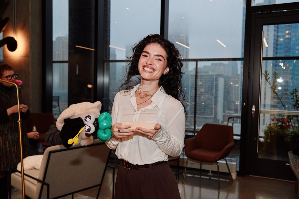 A smiling woman holding a clear rectangular award in front of a window overlooking a city.