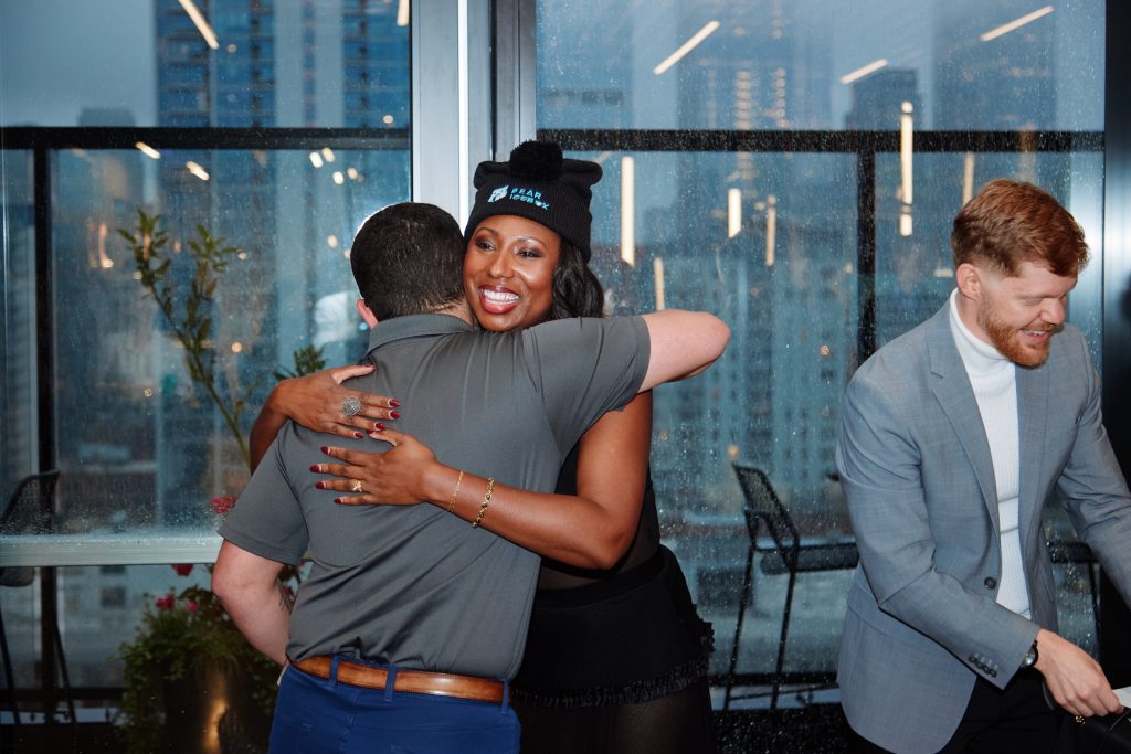A woman wearing a black beanie with the text “PEARLBOX” smiles while hugging a man, as a man in a grey suit jacket and white turtleneck looks on nearby.