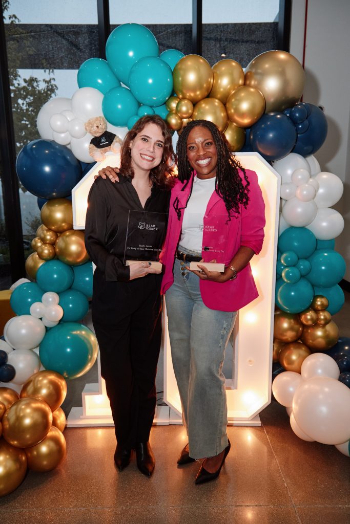Two women smiling and holding glass awards while standing in front of a large, illuminated number one sign decorated with blue, white, and gold balloons.