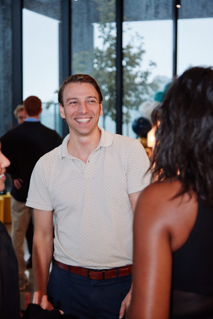 A man in a light-colored polo shirt smiles while talking to two people in a social setting.