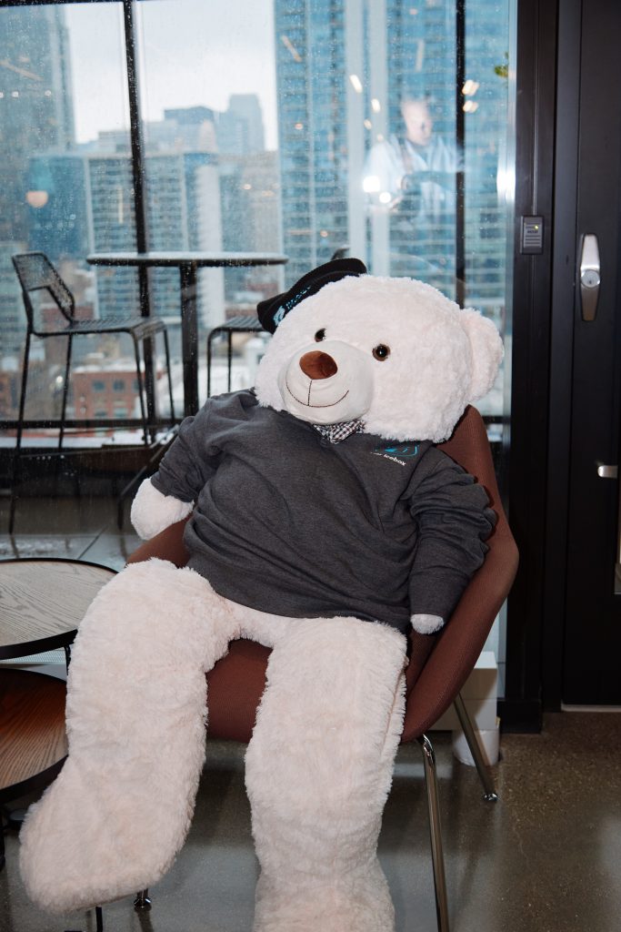 A large white teddy bear wearing a dark grey sweatshirt and a black beret sits in a chair in front of a window overlooking a city.